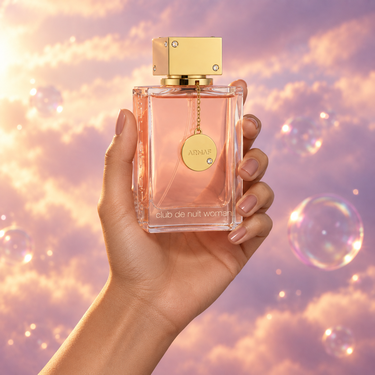 Una mano sostiene el frasco de Club de Nuit Woman de Armaf entre nubes rosadas.