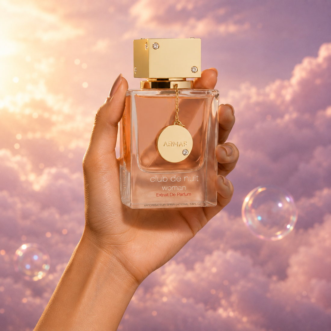 Una mano sostiene el frasco de Club de Nuit Woman Extrait de Parfum de Armaf entre nubes rosadas.