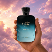 A hand holds the bottle of Hawas Malibu by Rasasi among pink clouds.