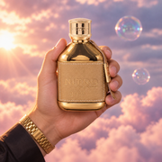 Una mano sostiene el frasco de Nitro Gold de Dumont entre nubes rosadas.