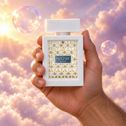 A hand holds the bottle of Rave Now White by Lattafa among pink clouds.