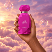 A hand holds the bottle of Uhud by Arabiyat Prestige among pink clouds.
